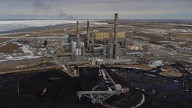 An industrial power plant with several smokestacks and large buildings is situated in a flat, expansive landscape. The surrounding area has patches of snow and water, with a network of roads and utility lines visible. An area of dark earth or coal is prominent in the foreground.