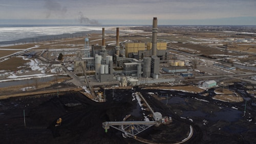 An industrial power plant with several smokestacks and large buildings is situated in a flat, expansive landscape. The surrounding area has patches of snow and water, with a network of roads and utility lines visible. An area of dark earth or coal is prominent in the foreground.