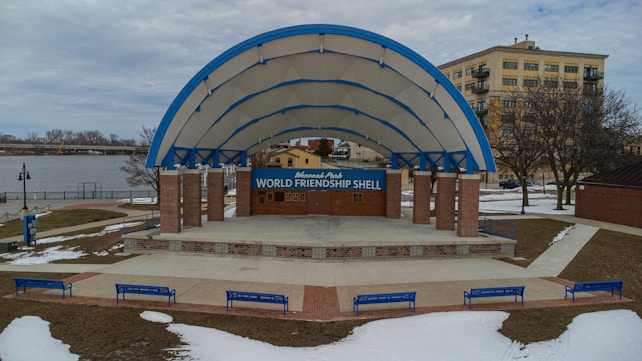 An outdoor amphitheater with a large, arched canopy labeled as the World Friendship Shell. The structure is supported by brick pillars and surrounded by a concrete sitting area. Blue benches are placed around the amphitheater, and patches of snow are visible on the ground. A river and a building are in the background.