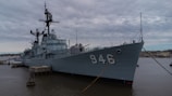 A large, grey military ship is docked at a pier with the number 946 prominently displayed on its side. The ship appears to have a complex array of structures and equipment on its deck, including antennas and radar systems. The sky is overcast, casting a muted light over the scene, and the water is calm, with a few distant structures visible on the horizon.