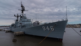 A large, grey military ship is docked at a pier with the number 946 prominently displayed on its side. The ship appears to have a complex array of structures and equipment on its deck, including antennas and radar systems. The sky is overcast, casting a muted light over the scene, and the water is calm, with a few distant structures visible on the horizon.