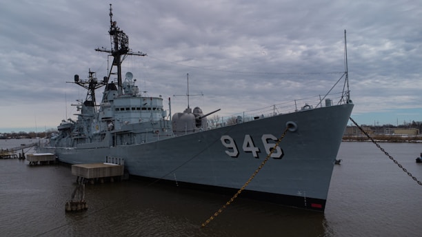 A large, grey military ship is docked at a pier with the number 946 prominently displayed on its side. The ship appears to have a complex array of structures and equipment on its deck, including antennas and radar systems. The sky is overcast, casting a muted light over the scene, and the water is calm, with a few distant structures visible on the horizon.