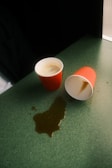 Two red paper cups are placed on a green surface, with one cup standing upright containing some liquid and the other is tipped over, causing a spill on the surface.