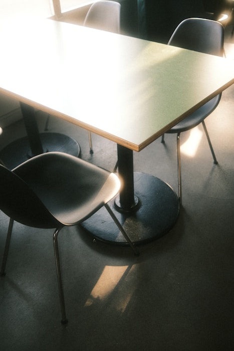 A minimalist dining setup featuring a rectangular table with a light surface and a black pedestal base. Surrounding the table are four modern, dark-colored chairs, with sunlight streaming in and casting soft shadows on the floor.
