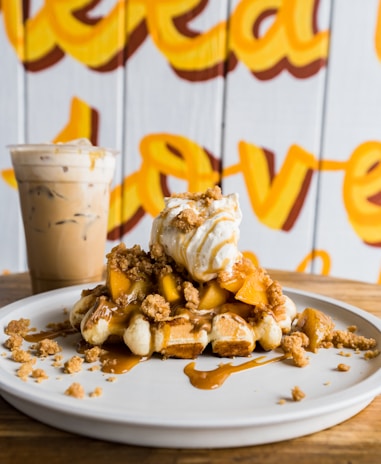 A delectable dessert featuring a waffle topped with ice cream, caramel sauce, and crumble. The plate sits on a wooden table with a cup of iced coffee in the background, against a vibrant wall with artistic yellow script.