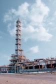 An industrial facility with a tall metal structure, resembling an oil refinery or processing plant. The structure is composed of steel beams, platforms, and numerous pipes. The sky is clear with a few scattered clouds, providing a backdrop to the facility.
