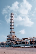 An industrial facility with a tall metal structure, resembling an oil refinery or processing plant. The structure is composed of steel beams, platforms, and numerous pipes. The sky is clear with a few scattered clouds, providing a backdrop to the facility.