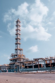 An industrial facility with a tall metal structure, resembling an oil refinery or processing plant. The structure is composed of steel beams, platforms, and numerous pipes. The sky is clear with a few scattered clouds, providing a backdrop to the facility.