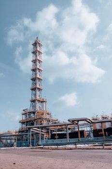 An industrial facility with a tall metal structure, resembling an oil refinery or processing plant. The structure is composed of steel beams, platforms, and numerous pipes. The sky is clear with a few scattered clouds, providing a backdrop to the facility.
