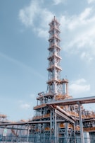 A large industrial structure with a tall metal tower featuring complex scaffolding and pipes. The sky is mostly clear with some clouds. The structure appears to be part of an oil refinery or chemical plant with visible walkways and ladders.
