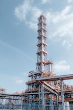 A large industrial structure with a tall metal tower featuring complex scaffolding and pipes. The sky is mostly clear with some clouds. The structure appears to be part of an oil refinery or chemical plant with visible walkways and ladders.
