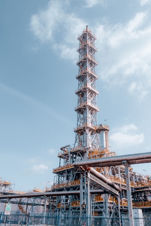 A large industrial structure with a tall metal tower featuring complex scaffolding and pipes. The sky is mostly clear with some clouds. The structure appears to be part of an oil refinery or chemical plant with visible walkways and ladders.