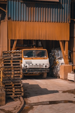 A white truck with an orange cab is parked inside an industrial building. The structure is made of corrugated metal sheets painted brown. Stacks of wooden pallets are on the left, and there is some machinery and large bags of materials on the right.