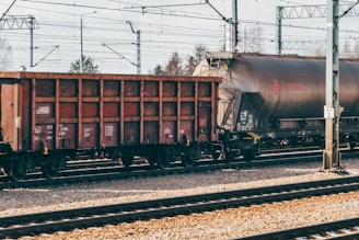 Freight train cars are stationed on railway tracks, with overhead electrical lines and pylons visible. The scene is clear, and the environment seems calm and industrial.