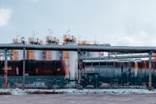 An industrial setting featuring large cylindrical storage tanks and freight train cars. The tanks are white with yellow access ladders and piping on top. The train cars have visible weathering, situated behind a metal fence. Overhead pipes run parallel to the tanks and ground.