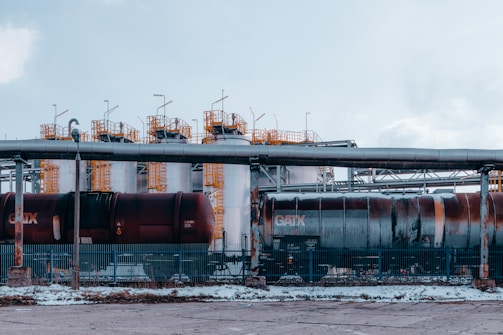 An industrial setting featuring large cylindrical storage tanks and freight train cars. The tanks are white with yellow access ladders and piping on top. The train cars have visible weathering, situated behind a metal fence. Overhead pipes run parallel to the tanks and ground.