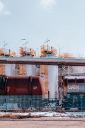 Large industrial storage tanks are situated behind a metal fence. Two rusty freight train cars are visible in front of the tanks. Metal structures with yellow railings are on top of the tanks, and the sky overhead is clear and blue.