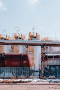 Large industrial storage tanks are situated behind a metal fence. Two rusty freight train cars are visible in front of the tanks. Metal structures with yellow railings are on top of the tanks, and the sky overhead is clear and blue.
