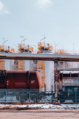 Large industrial storage tanks are situated behind a metal fence. Two rusty freight train cars are visible in front of the tanks. Metal structures with yellow railings are on top of the tanks, and the sky overhead is clear and blue.