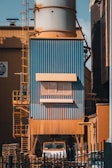 A large industrial structure with blue and orange metal cladding is visible, featuring a tall cylindrical tower. There is a ladder on the left side, and a truck is partially visible underneath. Pallets are stacked nearby, and the entire area is surrounded by a black fence.