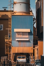 A large industrial structure with blue and orange metal cladding is visible, featuring a tall cylindrical tower. There is a ladder on the left side, and a truck is partially visible underneath. Pallets are stacked nearby, and the entire area is surrounded by a black fence.