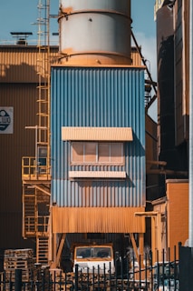 A large industrial structure with blue and orange metal cladding is visible, featuring a tall cylindrical tower. There is a ladder on the left side, and a truck is partially visible underneath. Pallets are stacked nearby, and the entire area is surrounded by a black fence.