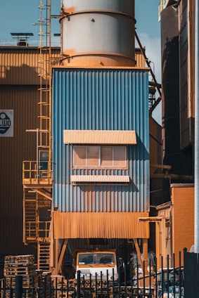 A large industrial structure with blue and orange metal cladding is visible, featuring a tall cylindrical tower. There is a ladder on the left side, and a truck is partially visible underneath. Pallets are stacked nearby, and the entire area is surrounded by a black fence.