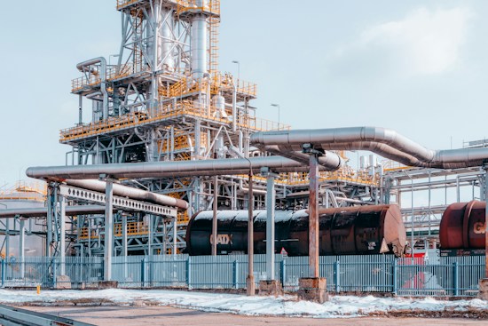 An industrial facility with a large complex of metal structures and pipelines. The scene depicts a network of interconnected pipes and cylindrical tanks, surrounded by a fence. The ground is partially covered in snow, suggesting a cold environment. The structures are primarily metallic with highlights of yellow safety railings.