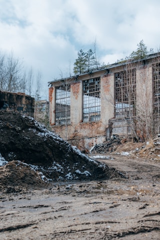 A misty abandoned industrial building surrounded by overgrown vegetation at dawn.