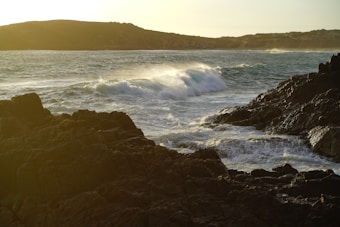 A rugged coastline with dark rocky cliffs is set against a powerful ocean. Waves crash dramatically against the rocks, spraying mist into the air. The sun hangs low, casting a golden glow across the scene, and the distant hills create a silhouette against the sky.