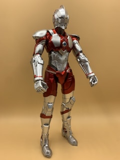 A detailed figure of a futuristic humanoid robot in armor, primarily featuring metallic silver and red colors. The design includes intricate details such as plating, joints, and a central chest component resembling an energy core. The figure stands upright against a plain tan background.