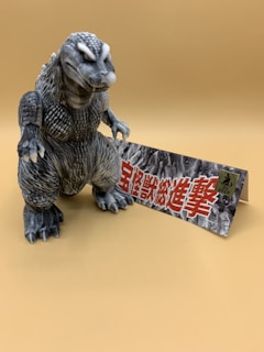 A gray, textured figure resembling a dinosaur or monster stands on a plain tan background, accompanied by a rectangular card with bold red Japanese characters and some smaller black text.