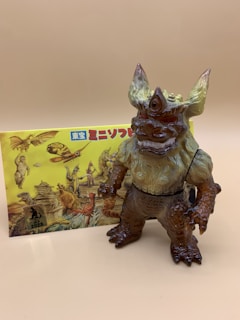 A colorful, detailed monster figurine with a single eye on its forehead stands prominently in front of a box featuring various illustrated creatures. The monster has large ears, sharp claws, and a textured body blending earthy tones. The background box has a vibrant design with multiple fantasy monsters in dynamic poses.