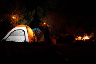 Two illuminated tents are set up in a dark outdoor setting surrounded by trees. A campfire is burning to the right, providing some additional light. The scene is dimly lit with warm, glowing tones from the fire and overhead lights.
