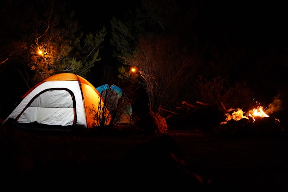 Two illuminated tents are set up in a dark outdoor setting surrounded by trees. A campfire is burning to the right, providing some additional light. The scene is dimly lit with warm, glowing tones from the fire and overhead lights.
