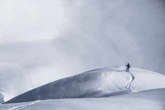 A lone skier makes their way up a vast, untouched snowy slope, leaving behind a single trail of ski tracks. The expansive snow-covered landscape stretches out into the distance, with subtle variations in texture and shadow highlighting the undulating terrain.