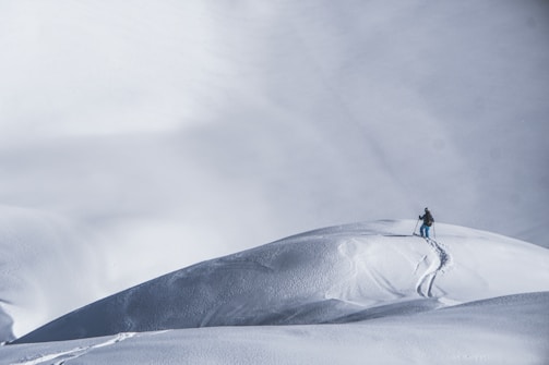 A lone skier makes their way up a vast, untouched snowy slope, leaving behind a single trail of ski tracks. The expansive snow-covered landscape stretches out into the distance, with subtle variations in texture and shadow highlighting the undulating terrain.