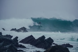 A surfer navigates a large, powerful wave near rocky shores, showcasing skill and balance. The water appears turbulent, with waves crashing against dark rocks.