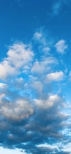 The image features a clear blue sky adorned with various fluffy white and gray clouds. The clouds range in size and shape, moving softly across the sky, suggesting a peaceful and calm weather condition.