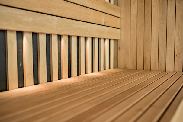 An interior of a wooden sauna featuring light wood panels and a bench. The space is lit by small recessed lights, casting a warm glow over the wood surfaces.
