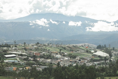 A scenic landscape featuring a village nestled among lush green hills with a background of majestic mountains. The sky is dotted with fluffy clouds, creating a serene and tranquil atmosphere. The village consists of small, colorful houses surrounded by trees and farmland.