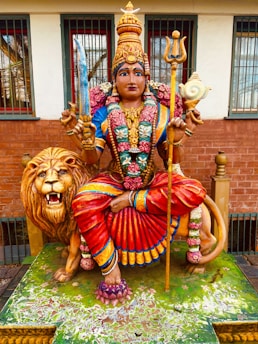 A colorful statue depicting a deity with an elaborate headdress, vibrant attire, and adorned with jewelry. The figure holds a sword in one hand and a trident in the other, sitting atop a majestic lion. The lion has a fierce expression, and the scene is set against a building with barred windows.