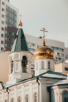 A beautiful church features ornate architecture with a striking green roof and a golden dome, adorned with Orthodox crosses on top. The building combines traditional elements with urban surroundings, as modern buildings are visible in the background.