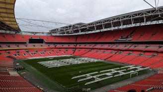 A large football stadium with red seats and a green field. The word 'Wembley' is visible in white letters on the seating section along the sideline. The stadium is equipped with a transparent dome-like roof structure supported by white metal frameworks. Several large, white lighting or maintenance rigs are positioned across the field.