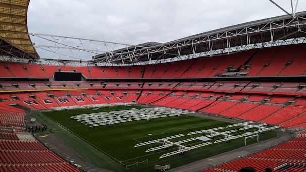 A large football stadium with red seats and a green field. The word 'Wembley' is visible in white letters on the seating section along the sideline. The stadium is equipped with a transparent dome-like roof structure supported by white metal frameworks. Several large, white lighting or maintenance rigs are positioned across the field.