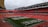 A large football stadium with red seats and a green field. The word 'Wembley' is visible in white letters on the seating section along the sideline. The stadium is equipped with a transparent dome-like roof structure supported by white metal frameworks. Several large, white lighting or maintenance rigs are positioned across the field.