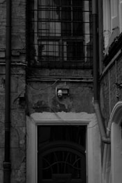 An old, weathered brick facade with a barred window on the upper section. A surveillance camera is mounted above a dark arched doorway. The walls show signs of deterioration, including cracks and peeling plaster. There are pipes and cables running along the wall.