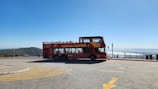 A bright red open-top double-decker bus is parked on a spacious paved area with scenic views in the background. The bus is adorned with vibrant graphics and has a sign that reads 'City Sightseeing Cape Town'. The area appears to be a viewpoint, surrounded by railings, with mountains and a clear blue sky in the distance. Several people can be seen sitting on the upper deck of the bus.