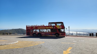 A bright red open-top double-decker bus is parked on a spacious paved area with scenic views in the background. The bus is adorned with vibrant graphics and has a sign that reads 'City Sightseeing Cape Town'. The area appears to be a viewpoint, surrounded by railings, with mountains and a clear blue sky in the distance. Several people can be seen sitting on the upper deck of the bus.
