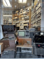 A vintage electronics store display behind a glass window, showcasing a collection of retro audio and video equipment. The shelves are filled with various devices, including old televisions, radios, amplifiers, and turntables. Reflection of the street and pedestrians create an overlay effect on the glass.